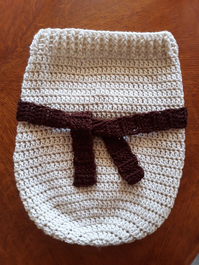 May include: A crocheted baby blanket in a cream color with a brown crocheted belt tied in a bow.