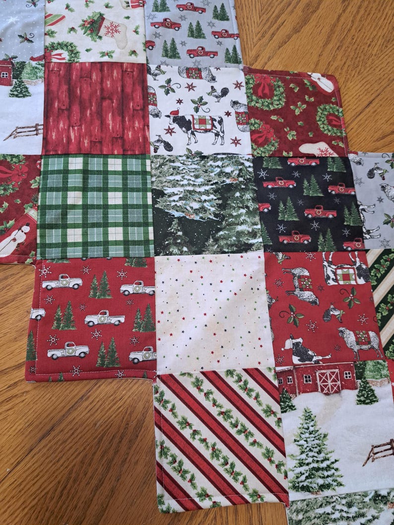 May include: A patchwork Christmas fabric with diverse patterns. The squares display red trucks, snowy scenes, plaid, and festive designs. The colour palette includes red, green, white, and grey.