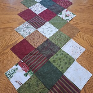 Pu&ograve; includere: Runner da tavolo trapuntato con motivo a rombi. Il runner presenta quadrati di vari tessuti nei toni del rosso, verde, marrone e bianco, con motivi tra cui strisce, aghi di pino e motivi natalizi. Il runner &egrave; posto su un tavolo di legno.