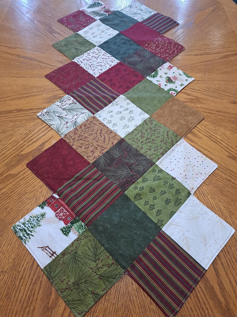 Pu&ograve; includere: Runner da tavolo trapuntato con motivo a rombi. Il runner presenta quadrati di vari tessuti nei toni del rosso, verde, marrone e bianco, con motivi tra cui strisce, aghi di pino e motivi natalizi. Il runner &egrave; posto su un tavolo di legno.