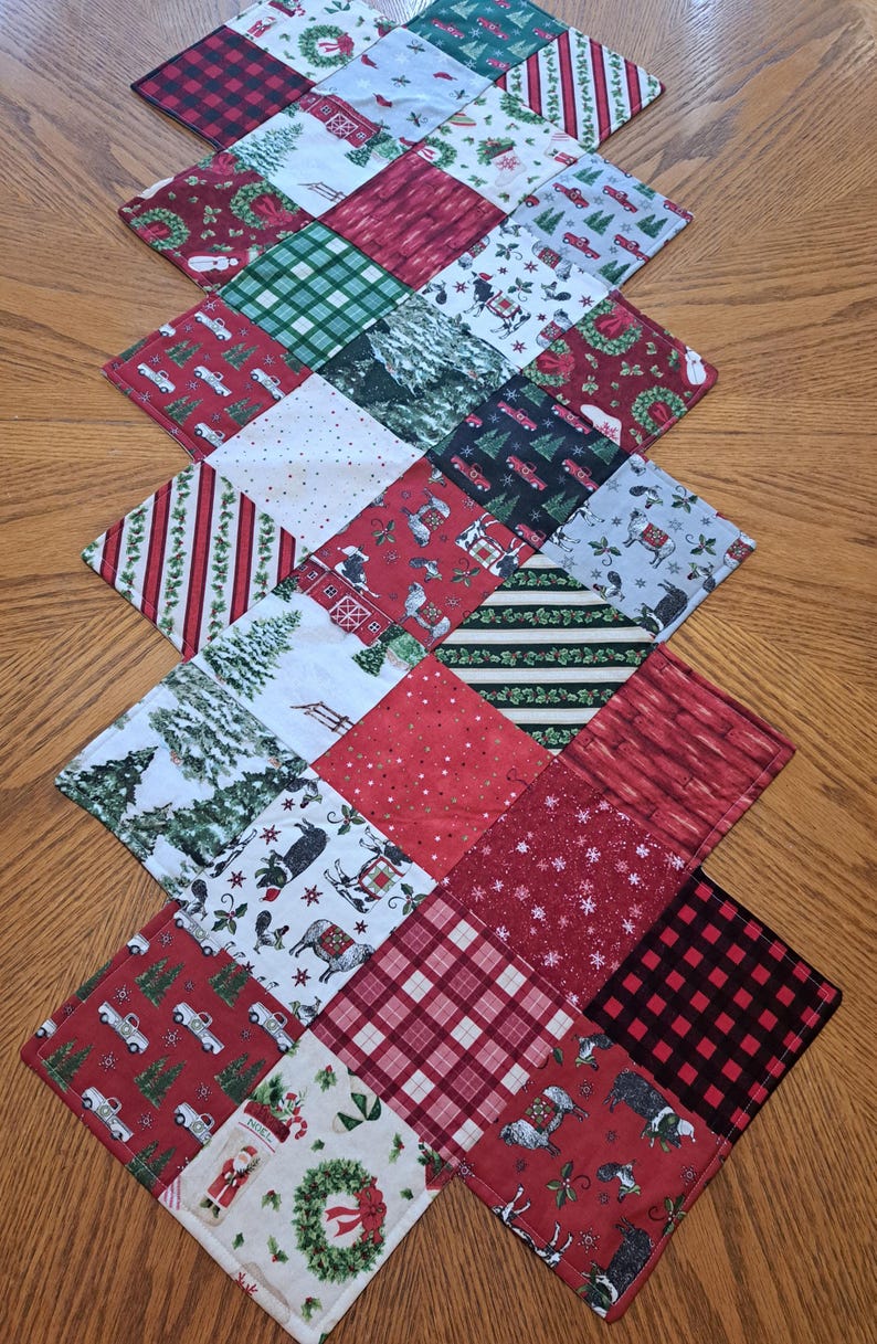 May include: A patchwork Christmas table runner with a diamond pattern. The runner displays festive designs such as red trucks, Christmas trees, plaid, and holiday-themed prints. The colour scheme includes red, green, white, and black.