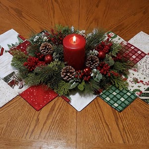 May include: A festive Christmas table runner with a central arrangement of a lit red candle, pine cones, and greenery. The runner is made of quilted fabric squares in red, green, and white holiday patterns. The table is made of wood.