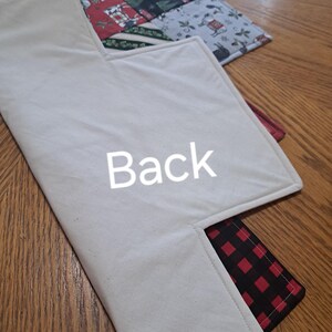 May include: A Christmas-themed quilted placemat with a cream-coloured back and a red and black plaid pocket. The front features a patchwork design with festive patterns and the word "Back" is printed on the back.