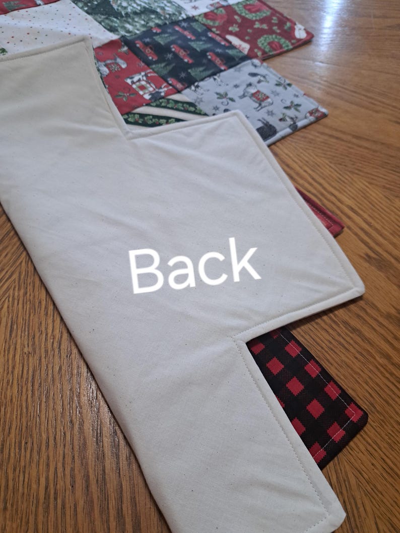 May include: A Christmas-themed quilted placemat with a cream-coloured back and a red and black plaid pocket. The front features a patchwork design with festive patterns and the word "Back" is printed on the back.