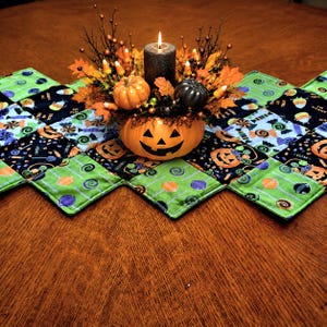 May include: A Halloween-themed table centerpiece featuring a carved pumpkin, a lit candle, and small pumpkins. The centerpiece sits on a quilted table runner with green and black diamond-shaped patches decorated with Halloween motifs.