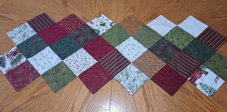 Pu&ograve; includere: Un runner da tavolo a forma di diamante con un design patchwork. I quadrati presentano vari tessuti a tema festivo in rosso, verde, bianco e marrone. I motivi includono strisce, motivi floreali e motivi di alberi di Natale. Il runner &egrave; posizionato su una superficie di legno.