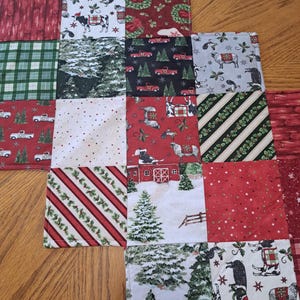May include: A patchwork Christmas table runner with squares of festive patterns. Designs include red trucks, cows, trees, and striped patterns in red, green, and white. The runner is placed on a wooden table.