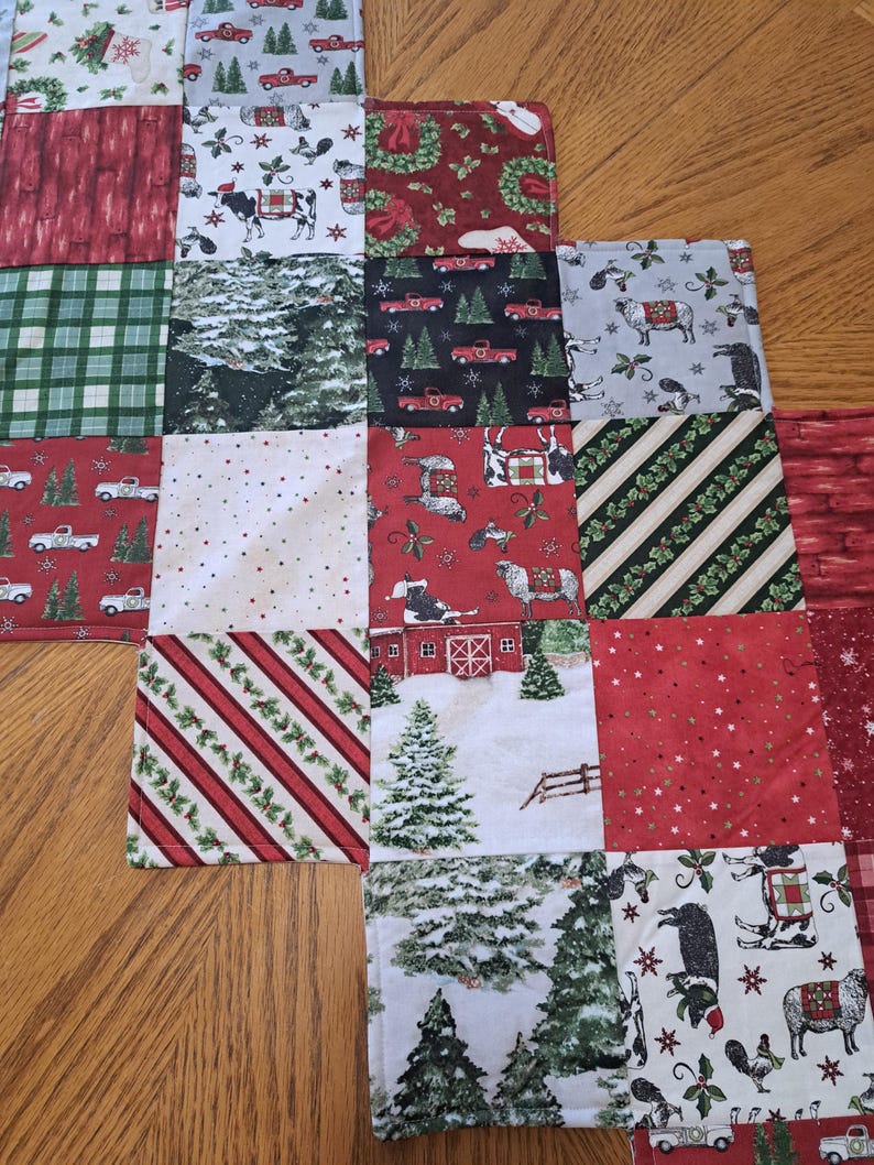 May include: A patchwork Christmas table runner with squares of festive patterns. Designs include red trucks, cows, trees, and striped patterns in red, green, and white. The runner is placed on a wooden table.