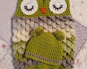 May include: A handmade crochet owl-shaped baby outfit in shades of green, gray, and white. The outfit includes a hooded blanket with large white eyes, black eyelashes, and an orange beak, plus a matching hat.