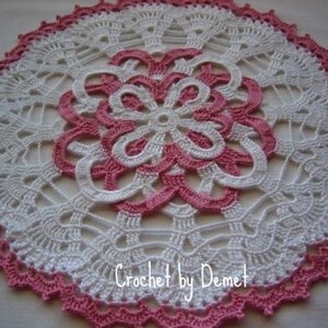 May include: A handmade, circular crochet doily in white and pink. The doily features a central floral design in pink, surrounded by white openwork. The outer edge is trimmed with a scalloped pink border. The text "Crochet by Demet" is visible.
