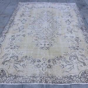 May include: A rectangular, beige-colored rug with an intricate floral pattern and a dark gray border. The rug's design features a central medallion and repeating floral motifs, creating a vintage aesthetic. The rug is placed on a gray tiled surface.