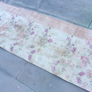 May include: Rectangular floral rug with a cream-colored central field and a light brown border. The rug features a pattern of pink and purple flowers with green leaves, creating a vintage aesthetic. The rug is placed on a gray stone surface.