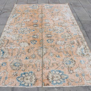 May include: A rectangular, vintage-style rug with a peach-colored background and a floral pattern in shades of blue, cream, and brown. The rug's design features stylized flowers and leaves, creating a classic, aged appearance. The rug is placed on a gray stone surface.