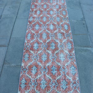 May include: A long, rectangular runner rug featuring a repeating damask-style pattern in faded terracotta orange and muted light blue. The textile has a weathered appearance with visible fringe at both ends and a symmetrical, intricate floral motif.