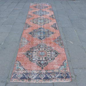 Peut inclure: Un long tapis rectangulaire avec un motif complexe. Le tapis présente un fond de couleur corail avec des motifs géométriques ornés bleu foncé et beige. Les bords du tapis sont finis avec une bordure bleu foncé. Le tapis est posé sur une surface carrelée grise.