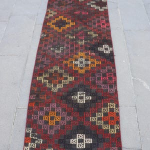 May include: A long, rectangular rug with a geometric pattern in shades of red, black, brown, orange, and white. The rug features a repeating design of squares and diamond shapes, creating a textured appearance. The rug is likely made of woven material.
