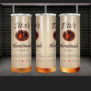Peut inclure: Trois gobelets en acier inoxydable avec un fond cr&egrave;me et un logo Tito's Vodka noir et orange. Les gobelets ont une bordure dor&eacute;e et indiquent "Vodka artisanale" et "Austin, Texas".