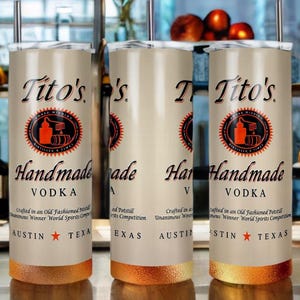 Peut inclure: Trois gobelets en acier inoxydable avec un fond de couleur cuivre. Les gobelets sont d&eacute;cor&eacute;s d'un fond blanc et d'un texte noir qui dit "Tito's Handmade Vodka". Le texte est entour&eacute; d'un cercle rouge avec un contour noir. Les gobelets sont tous de la m&ecirc;me taille et de la m&ecirc;me forme.