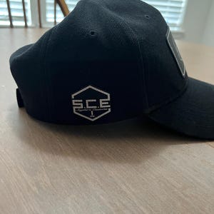May include: A black baseball cap featuring a silver hexagonal patch on the side. The patch displays the text S.C.E. and the words Snyder's Customs underneath. The cap has a structured crown, a curved brim, and a small button at the top center.