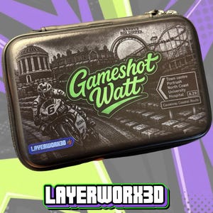 May include: A rectangular black zippered carrying case featuring a graphic print of a motorcycle racer, a roller coaster labelled Barrys Big Dipper, and a road sign listing Town centre, Portrush, North Coast, University, Hospital, A29, and Causeway Coastal Route. The text Gameshot Watt is printed in green, and the logo Layerwork3d is visible.