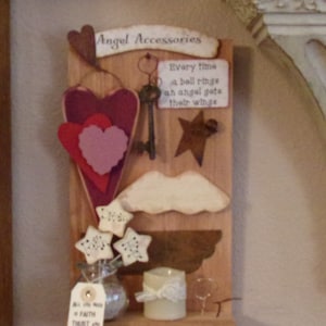 Puede incluir: Un letrero de madera con el texto "Angel Accessories" y la cita "Every time a bell rings an angel gets their wings". El letrero está decorado con corazones, estrellas, una llave y un ala blanca.