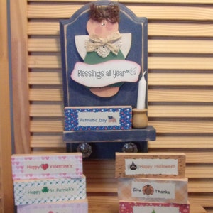 May include: A wooden shelf with a blue painted angel figurine and a candle. The shelf is decorated with small wooden blocks with different holiday themes, including "Happy Valentine's", "Happy St. Patrick's", "Happy Easter", "Happy Halloween", "Give Thanks", "Happy Holidays", and "Happy Birthday".