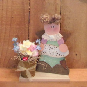 May include: Wooden figurine of an angel holding a sign that says "Bless My Garden". The angel is wearing a purple dress with a white lace collar and has brown hair. The angel is standing on a wooden base with a small pot of flowers in front of it.