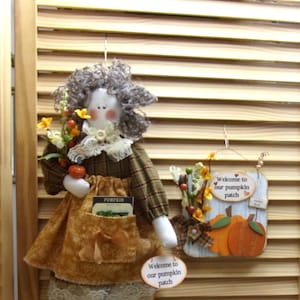 May include: A fall-themed decorative display featuring a doll with a plaid dress, holding flowers and a pumpkin. Includes signs that say "Welcome to our pumpkin patch" and a small pumpkin patch scene. The doll has a pocket with a seed packet that says "PUMPKIN".