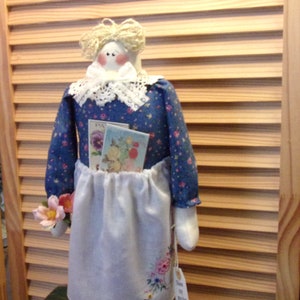 May include: A handmade doll wearing a blue dress with white lace trim and a white apron. The doll is holding seed packets and a small bouquet of flowers. The doll is standing on a white doily on a green surface. The tag on the doll says "Seeds for the Garden".