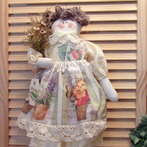 May include: A handmade rag doll with brown hair, wearing a white dress with a lace trim and a floral print. The doll is holding a bouquet of flowers and a packet of seeds that says "Sweet Pea".