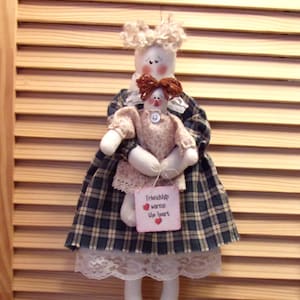 May include: A handmade doll wearing a plaid skirt with lace trim, holding a smaller doll. The larger doll has a sign that reads "Friendship warms the heart".