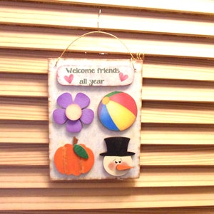 May include: Wooden sign with the text "Welcome friends all year" and four decorative elements: a purple flower, a beach ball, a pumpkin, and a snowman.