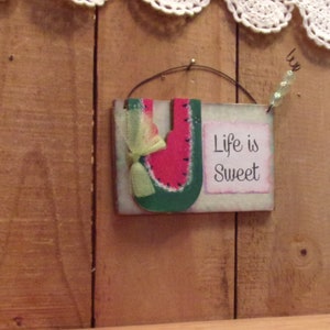 Può includere: Un cartello di legno con un ritaglio di anguria e il testo "Life is Sweet" dipinto su di esso. Il cartello è appeso a un filo e ha un fiocco verde sull'anguria.