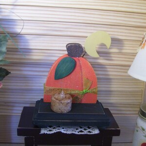 May include: A wooden pumpkin-shaped candle holder with a crescent moon design and a small candle. The holder is painted orange with green and gold accents. It is sitting on a dark brown wooden table.