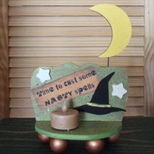 Op de afbeelding: Een groene houten kaarsenhouder met een heksenthema, met een zwarte heksenhoed, een halve maan en een bordje met de tekst "Time to cast some nasty spells". De houder heeft een kleine kaars in het midden en staat op vier ronde poten.