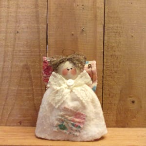 Könnte beinhalten: Eine handgemachte Stoffpuppe Engel mit einem weißen Kleid, braunen Haaren und einem rosa-weißen Patchworkrock. Der Engel hat eine Knopfnase und bemalte Augen.