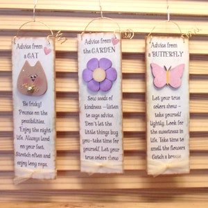 May include: Three wooden signs with inspirational quotes. The first sign features a cat and the quote "Be frisky! Pounce on the possibilities. Enjoy the night life. Always land on your feet. Stretch often and enjoy long naps.". The second sign features a purple flower and the quote "Sow seeds of kindness - listen to sage advice. Don't let the little things bug you - take time for yourself. Let your true colors show.". The third sign features a butterfly and the quote "Let your true colors show - take yourself lightly. Look for the sweetness in life. Take time to smell the flowers. Catch a breeze."