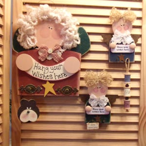May include: Three wooden angel ornaments with painted faces and curly hair. Each angel holds a sign that says "Hang your wishes here". The angels are decorated with lace, stars, and other embellishments.