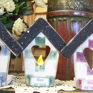 May include: Three wooden birdhouse-shaped candle holders with black roofs and white bases. Each house has a heart-shaped cutout and is decorated with a different plaid pattern in blue and white, green and white, and purple and white. Each house has a sign that reads "Bless our nest".