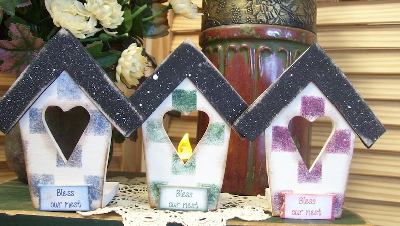 May include: Three wooden birdhouse-shaped candle holders with black roofs and white bases. Each house has a heart-shaped cutout and is decorated with a different plaid pattern in blue and white, green and white, and purple and white. Each house has a sign that reads "Bless our nest".