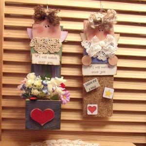 May include: Two wooden angel figurines with painted faces and lace trim. The angel on the left has a flower pot attached to its base. The angel on the right has a corkboard attached to its base. Both angels have a heart and the text "I will watch over you".