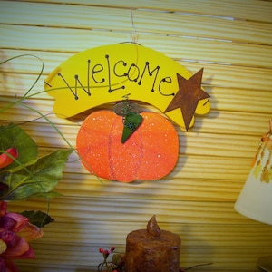 May include: A wooden welcome sign with a yellow background and black lettering. The sign has a pumpkin and a star attached to it. The sign is hanging on a wooden slat wall.
