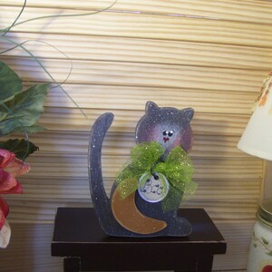 May include: A wooden black cat figurine with a brown crescent moon shape and a green bow. The cat has a tag that says "Boo". The cat is on a small black table. A lamp with a white shade is to the right.