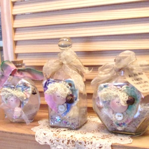 May include: Three glass bottles filled with colorful fabric scraps, beads, and glitter. Each bottle has a different shaped top and is decorated with a ribbon and a small doll-like figure.