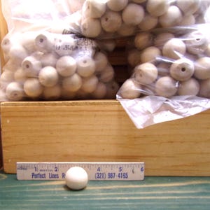 May include: Wooden craft beads in clear plastic bags, stacked on a wooden box. A single bead rests on a green surface next to a ruler. The ruler has the text "Perfect Lines R" and a phone number.