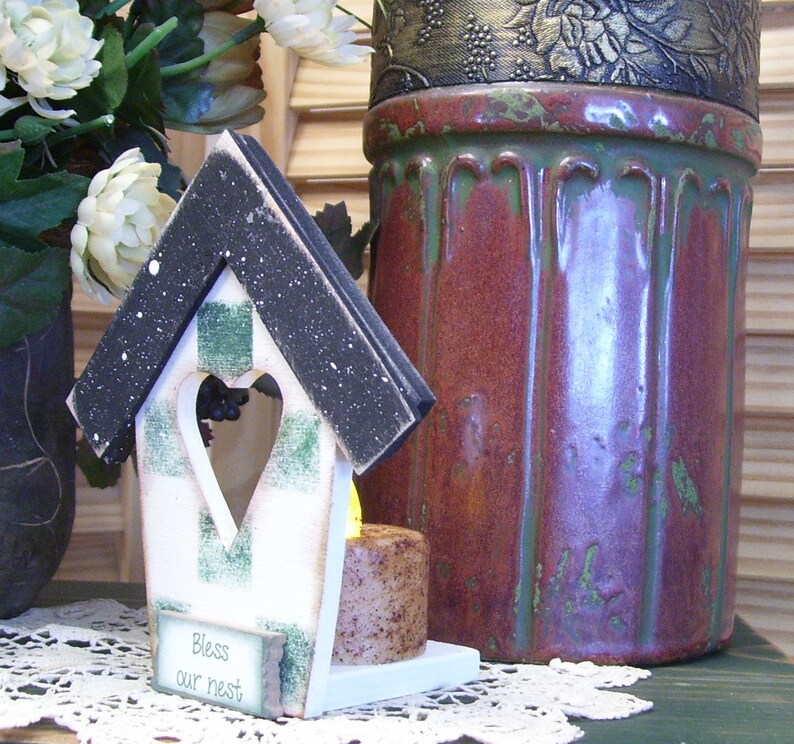 May include: A white birdhouse with a black roof and green and white checkered accents. The birdhouse has a heart-shaped opening and a sign that reads "Bless our nest".