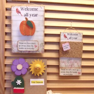 May include: Three wooden signs with a welcome theme. The top sign features a red cardinal, an orange pumpkin, and the text "Welcome all year". The middle sign is a corkboard with a red cardinal and the text "Welcome all year". The bottom sign features a purple flower, a yellow sun, a green mitten with a snowflake, and the text "Four Seasons".