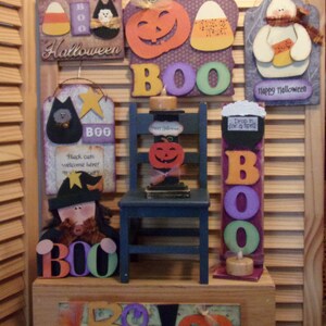 Op de afbeelding: Een verzameling Halloween-decoraties met verschillende houten borden met de woorden "Halloween", "BOO" en "Drop in for a spell". Er zijn ook pompoenen, geesten en een heks met een zwarte hoed en een bezem.