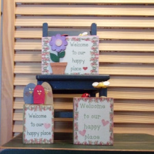 May include: Three wooden signs with a floral design and the text "Welcome to our happy place". The signs are sitting on a small wooden chair.