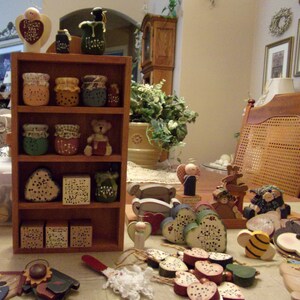 May include: A wooden shelf unit with various handmade wooden ornaments and figurines. The shelf is filled with jars, angels, bears, and hearts. The shelf is on a table with a white tablecloth. There are more ornaments on the table.